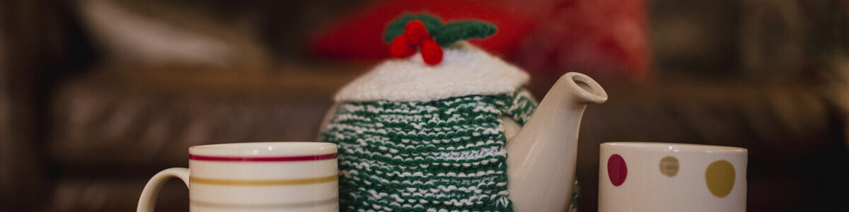 A tea pot covered in a green and white knit, two mugs, a gold bauble and a plate of mince pies.