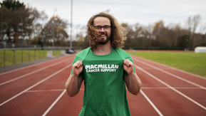 A man standing on a running track, proudly holds out the Macmillan logo on his running top to show he's running for Macmillan Cancer Support.