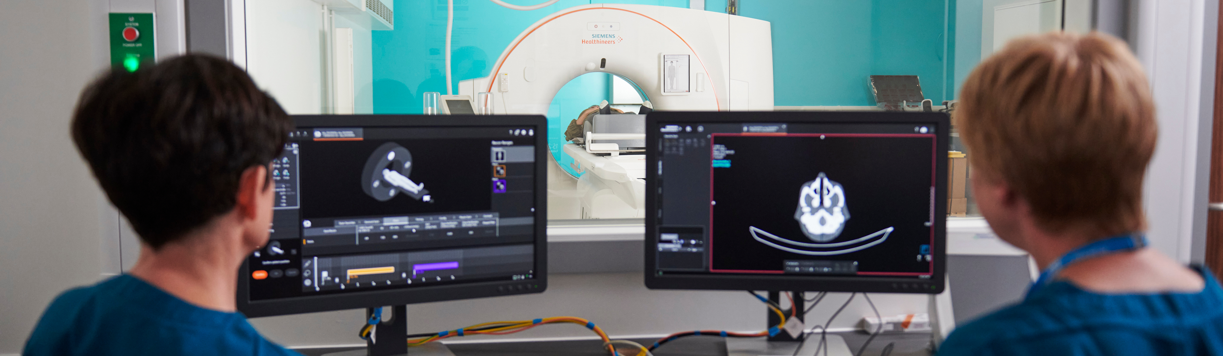 An image of two people looking at scans on a computer. Behind the screens is a window showing an MRI machine. 