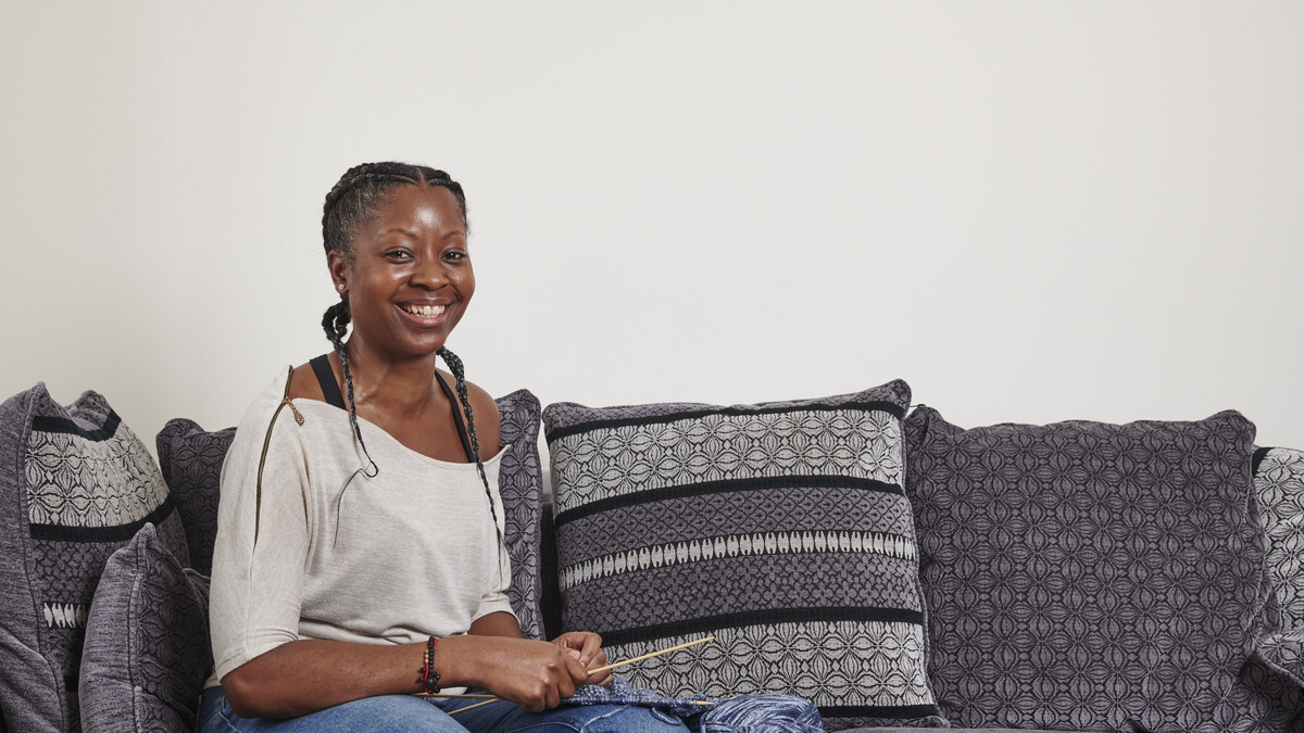 A photo of Bei, who was diagnosed with colorectal cancer in December 2014. In the image she is sitting on a sofa, smiling at the camera and has knitting materials in her hands.