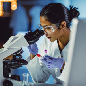 A woman in a lab looking into a microscope, holding two small blood test tubes in her left hand. 