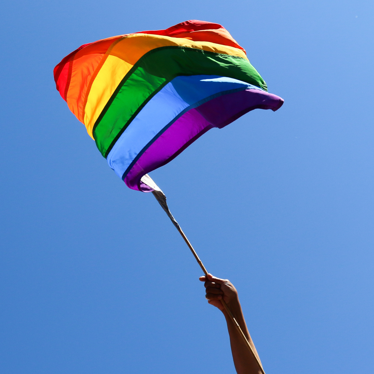 Two rainbow flags being waved against the backdrop of a blue sky.