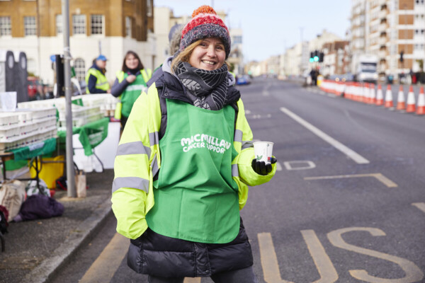 A volunteer stands on a closed off road at the Brighton Half Marathon 2023. They are wearing a Macmillan branded bib/vest and holding a cup of water to offer to runners.
