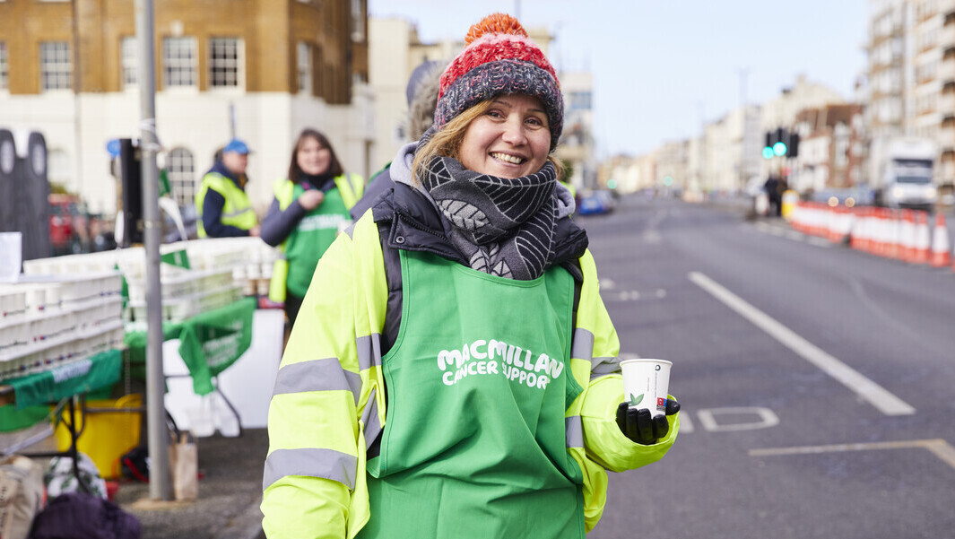 A volunteer stands on a closed off road at the Brighton Half Marathon 2023. They are wearing a Macmillan branded bib/vest and holding a cup of water to offer to runners.