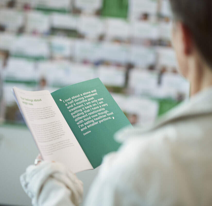 A patient reads a booklet produced by Macmillan in a Macmillan Support Centre. In the background you can see a shelf of other booklets. 