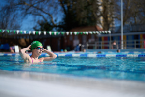 A person in a pool. They are wearing swimming goggles and a green Macmillan swimming cap. 