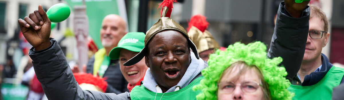 A person is standing and cheering outside at the London Landmarks Half Marathon. They're wearing a green Macmillan top and holding a green maraca in their left hand. On their head is a gold hat with red tassles. 