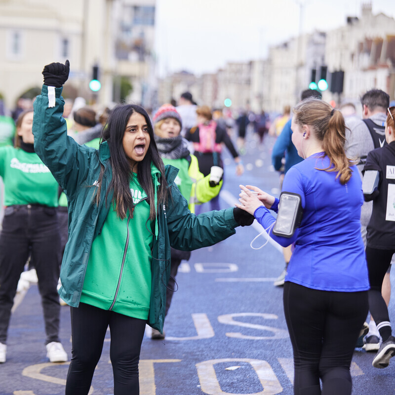 A woman is standing outside on a road. She is cheering on runners who pass by and is handing one a cup of water.