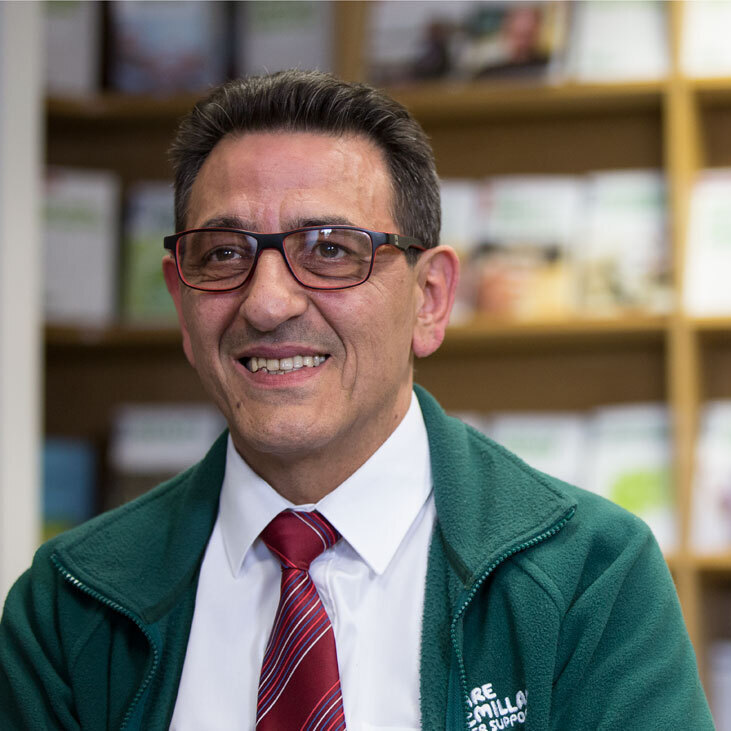 Mario, a middle-aged man wearing glasses, is sitting in front of shelves with cancer information booklets.