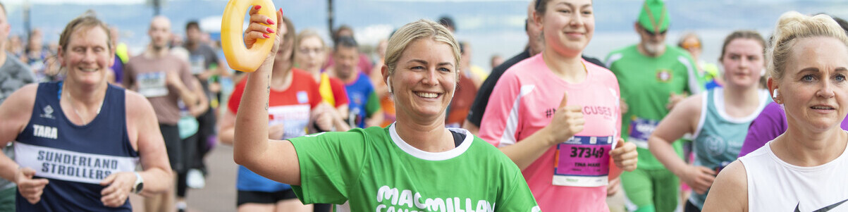 A person is running in the Edinburgh marathon. They are wearing a green Macmillan running top. In their hand they are holding up a yellow water bottle. 