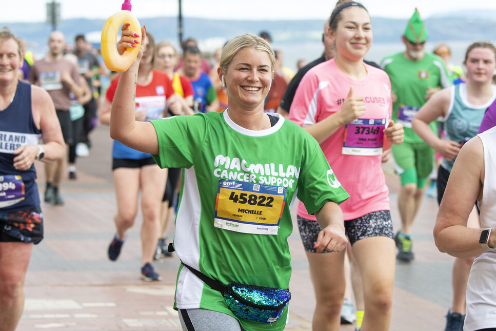 A person is running in the Edinburgh marathon. They are wearing a green Macmillan running top. In their hand they are holding up a yellow water bottle. 