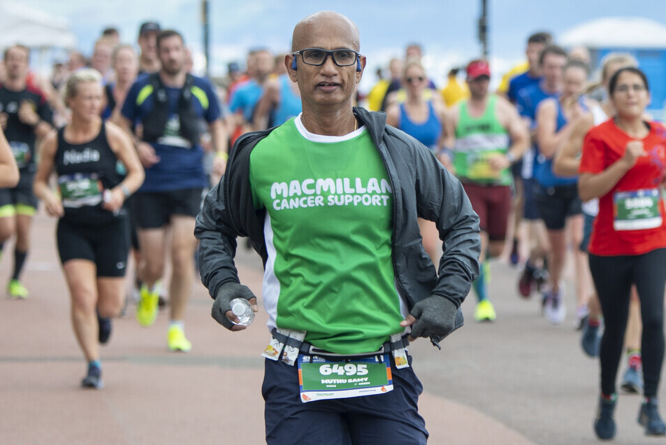 The runner is wearing a green Macmillan running top. They are also wearing a grey jacket and gloves, and blue shorts. Behind them are others runners. 