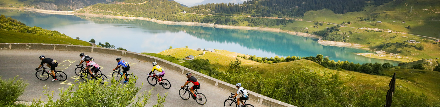 Group of 10 cyclists cycling up a hill with a lake and mountains in the background.