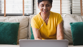 A person wearing a yellow t-shirt working on a laptop. They have white earphones on and they are sitting on a sofa in front of a window.