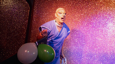 A person on stage wearing makeup, a wig cap and surgical gown