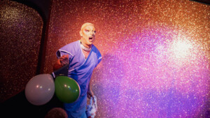 A person on stage wearing makeup, a wig cap and surgical gown