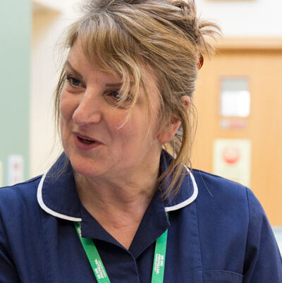 Shez, wearing her dark blue nurse's uniform and a green Macmillan lanyard, is talking to another nurse. They are in discussion.