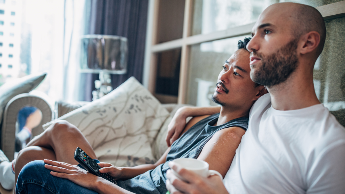 Two men sitting on a sofa watching tv. One has his arm around the other's shoulder.