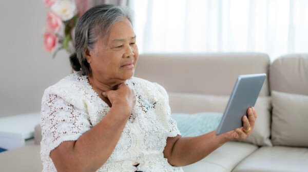 A woman is sat down. She is holding up a tablet. She is wearing a white top and has her other hand near her throat.