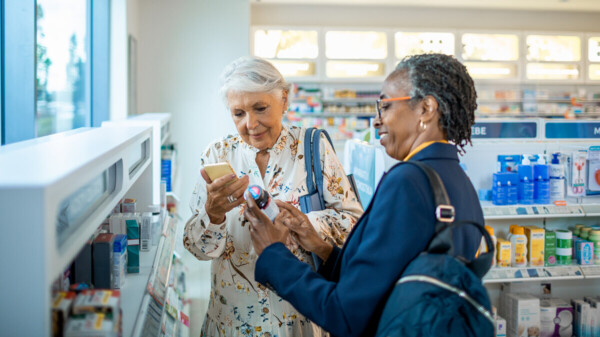A close up of two women browsing the shelves at a pharmacy. The person on the left is wearing a white blouse and the person on the right is wearing a blue blazer. 
