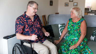 Jane Boulton (pictured right) is a Macmillan Social Care Coordinator. She is sat on a sofa listening to a person in a wheelchair (pictured left) speak. Jane is wearing a bright green patterned dress. The person on the left is wearing a blue and pink patterned shirt and tan trousers. 
