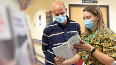 Emily (pictured right) is a Macmillan Cancer Information and Support Assistant. She is wearing a yellow patterned dress. Emily is speaking to a patient who is on her right. They are wearing a long sleeved blue t-shirt. 