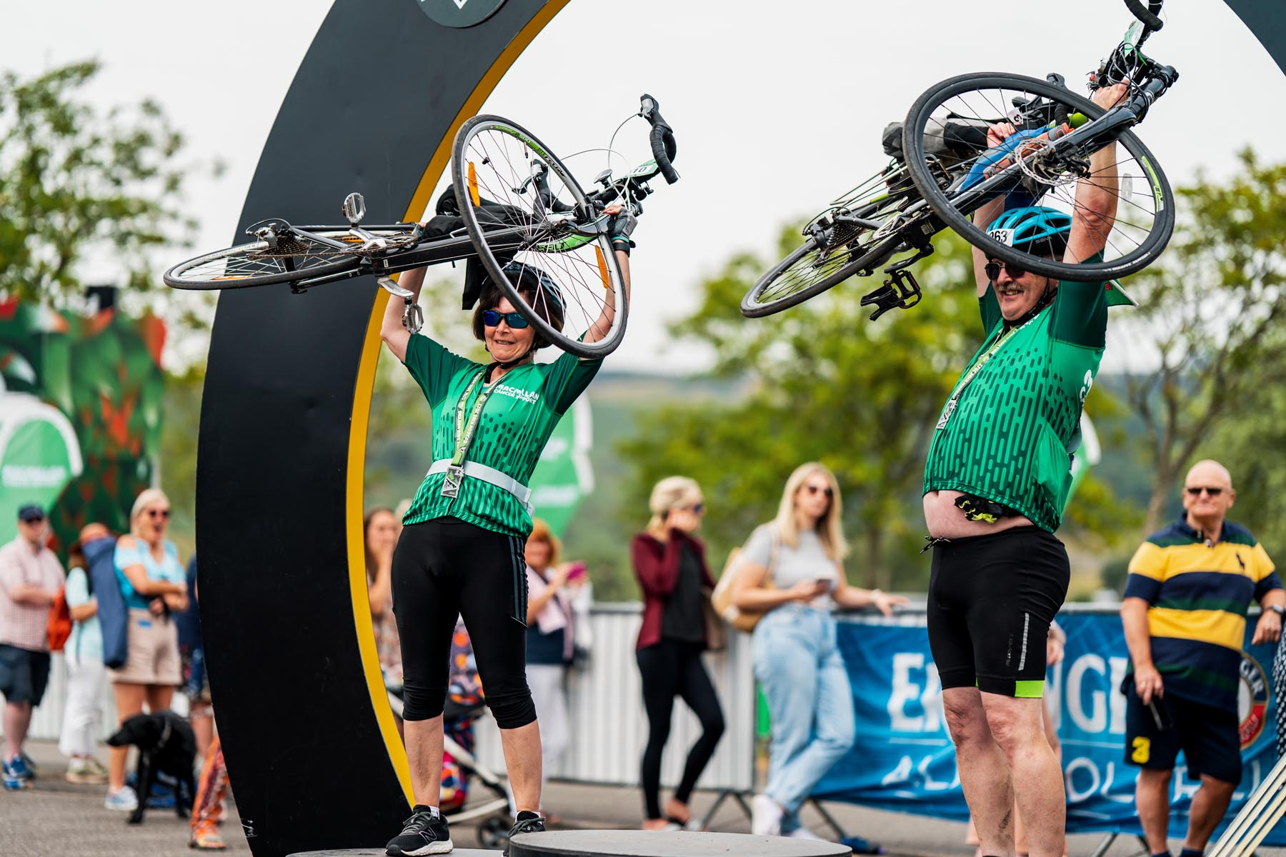 Two people in green Macmillan cycling tops standing at finish line. Both holding their bicycles in their arms above their heads and smiling. 