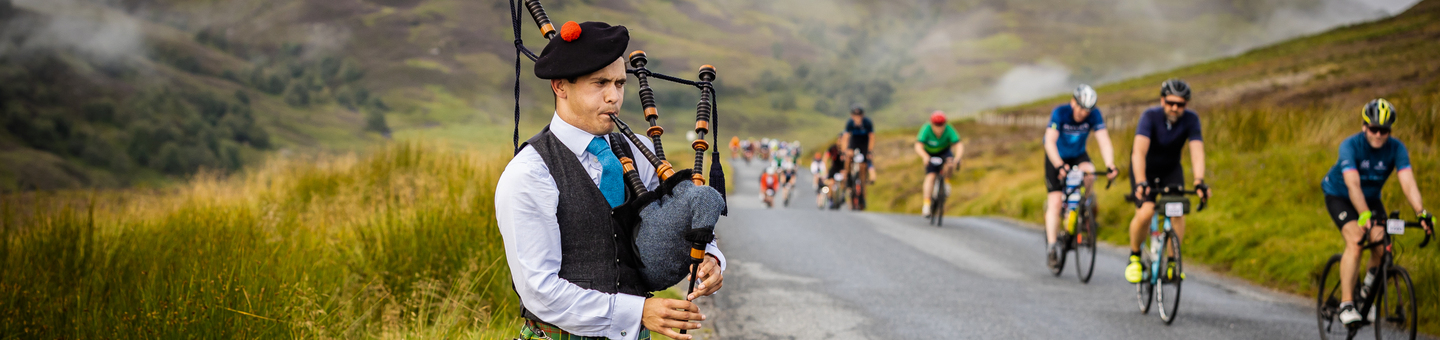 A man playing the bagpipes by the side of a mountain road beside cyclists.