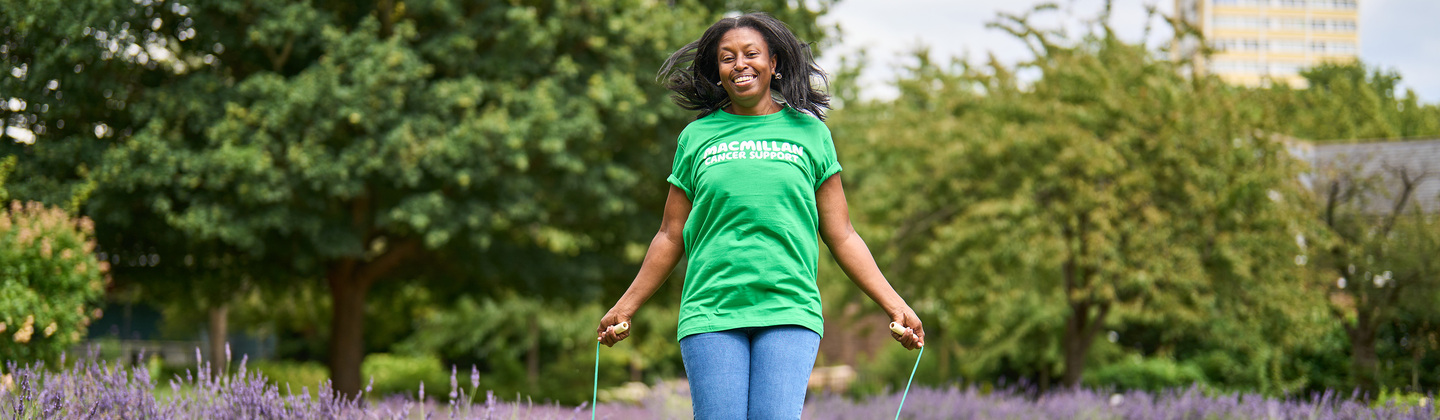 A woman is outside and is skipping a rope in a field. She is wearing a green Macmillan top, jeans, and white trainers. The rope she is skipping is green and behind her is lavender plants.
