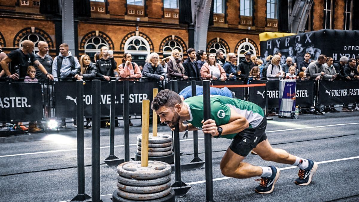 A person competing at the HYROX Birmingham event. They are hunched over pushing a contraption that has weights on it. 