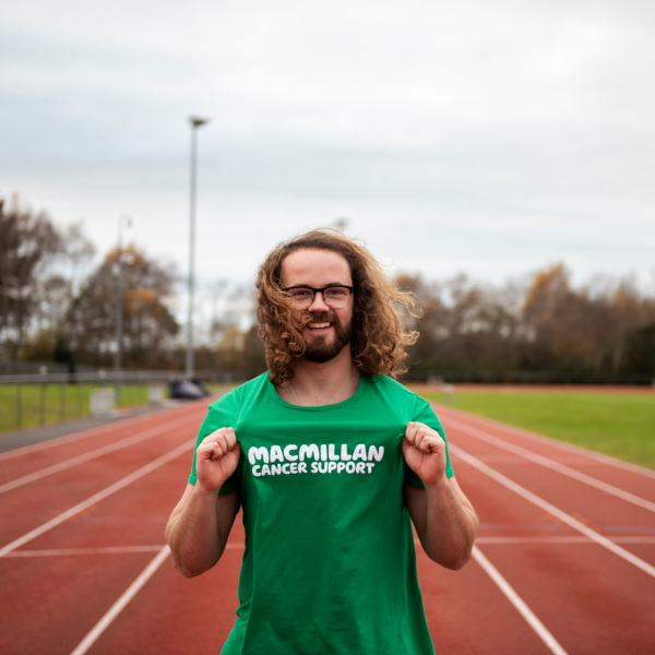 A man standing on a running track, proudly holds out the Macmillan logo on his running top to show he's running for Macmillan Cancer Support.