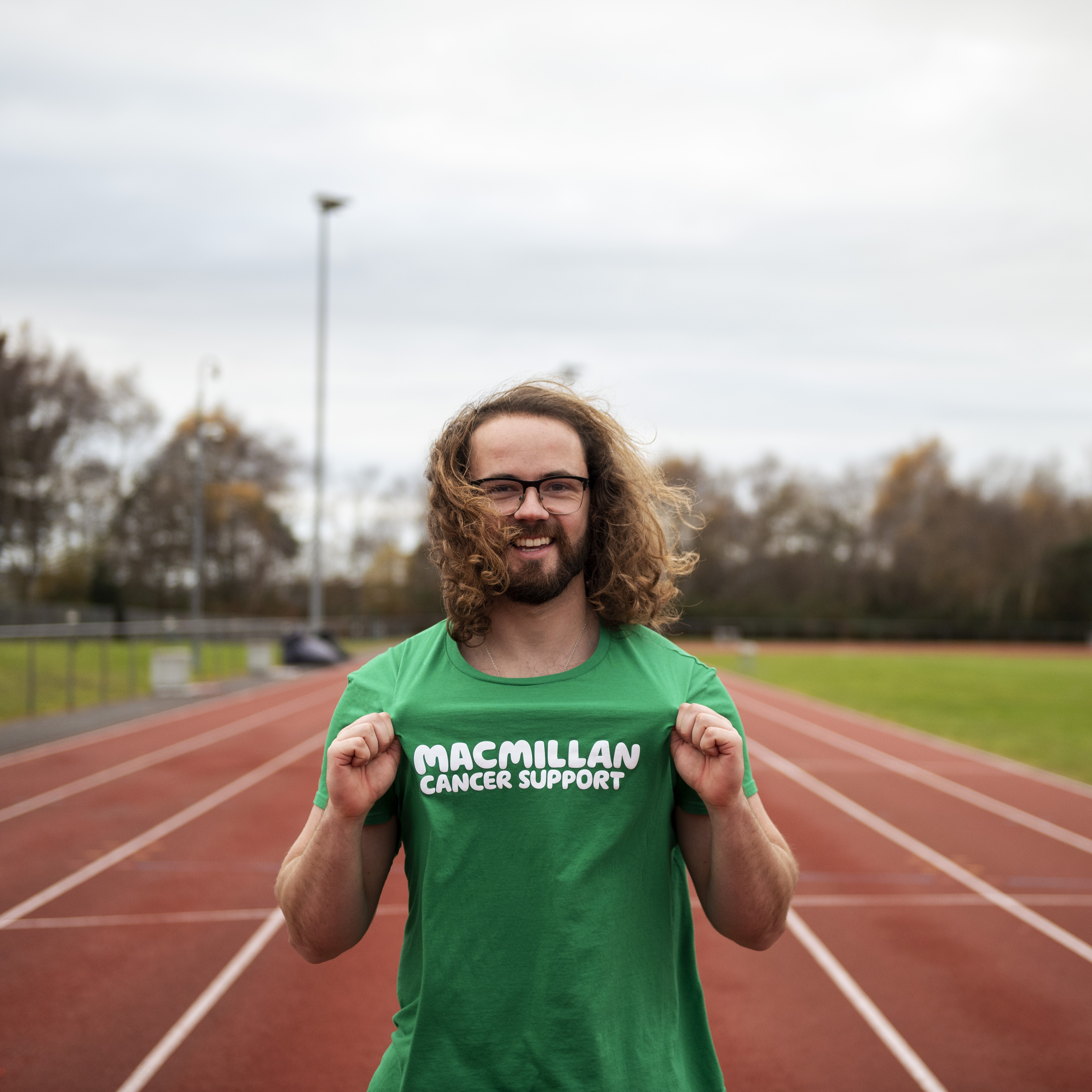 A man standing on a running track, proudly holds out the Macmillan logo on his running top to show he's running for Macmillan Cancer Support.