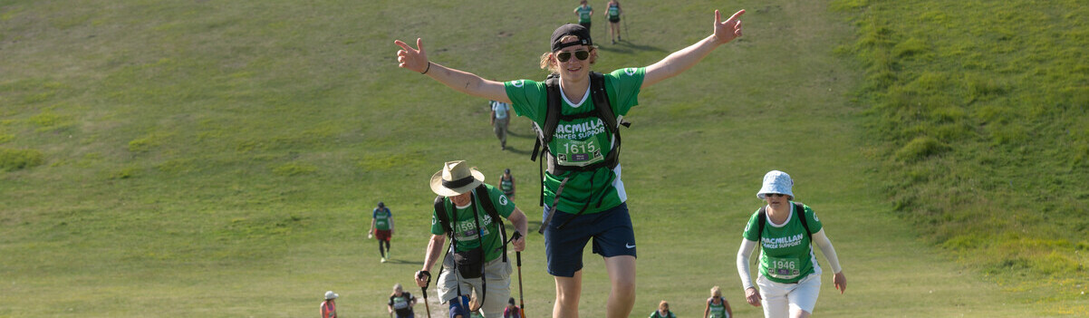 A person in a green Macmillan top is looking at the camera. Their arms are in the air and pointed outwards. Behind them are other people who are walking up a hill. 
