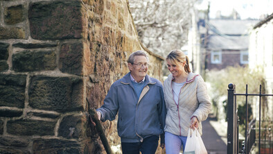 A pair appear outside and walking up a flight of stairs. The older person on the left is wearing a blue jumper and a blue jacket. The person on the right is wearing a white top and white jacket. They are also carrying a white shopping bag. 