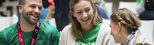 Three people are sitting down and talking. The person on the left is wearing a Macmillan running top and a metal from the London Marathon. The person in the middle is wearing a Macmillan top and the person on the right is wearing a trench coat.