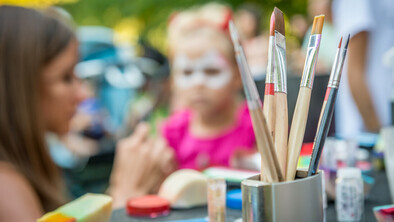 In the forefront of the image is a cup with paint brushes in it. In the background is a blurred view of a person face painting a younger person.