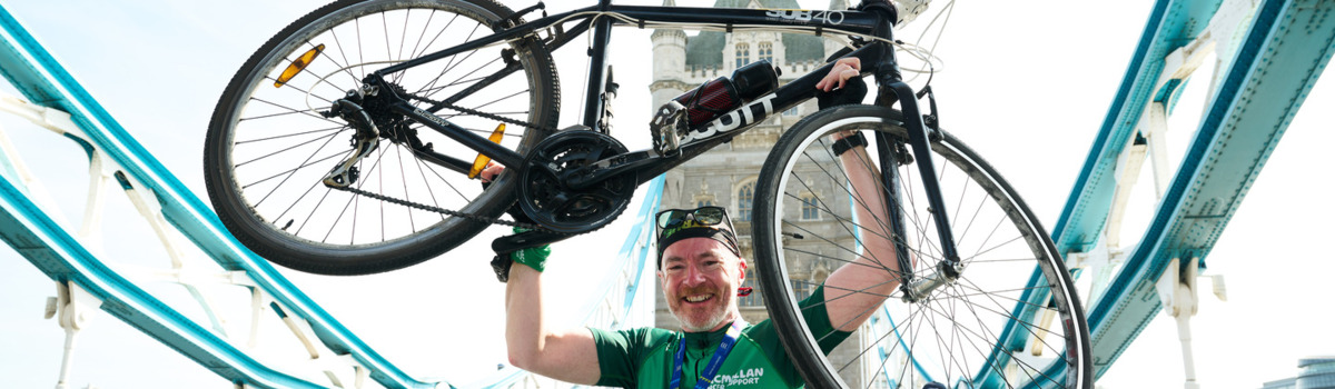 A cyclist in Macmillan top standing on the Tower Bridge in London holding a bicycle in both arms above their head. 