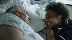 A person is resting in a hospital bed. They are laying on their side. Next to them, a nurse is leaning her arms and head on the edge of the bed. They appear to be talking to the patient in the hospital bed.