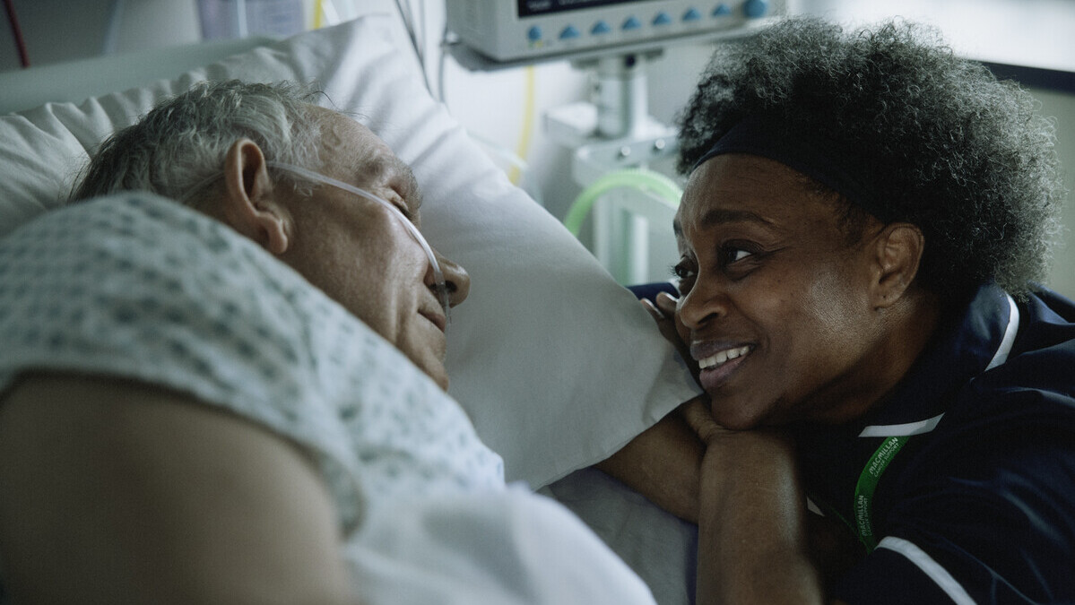 A person is resting in a hospital bed. They are laying on their side. Next to them, a nurse is leaning her arms and head on the edge of the bed. They appear to be talking to the patient in the hospital bed.