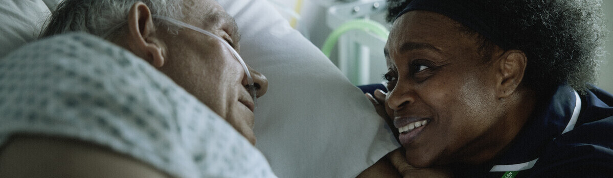 A person is resting in a hospital bed. They are laying on their side. Next to them, a nurse is leaning her arms and head on the edge of the bed. They appear to be talking to the patient in the hospital bed.