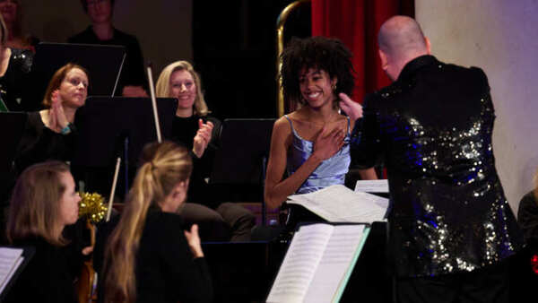 A woman is standing on a stage in a blue gown. She has a hand on her chest and is smiling. Around her are orchestra performers and the conductor and they are applauding the woman. 