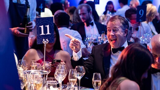 A man is smiling and pointing at a table number. He is in black tie and there are others sitting at his table and at tables around them.