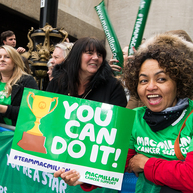 A Macmillan cheer point at the London Marathon. A person in a green Macmillan t shirt is holding up a poster that says 'You can do it!' with a picture of a trophy on it. There is a group of people standing behind a barrier holding up green Macmillan banners.
