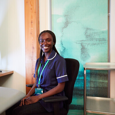 A nurse is sat in an office chair and facing towards the camera smiling. She has long braids in her hair, is wearing a blue nurse's uniform, and has on a green lanyard. Next to her is a desk with a computer. 