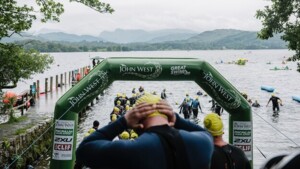 A person is standing with their hands on the back of their head. They have a yellow swimming cap on and a wetsuit. In front of them people in similar clothing are entering a body of water. 
