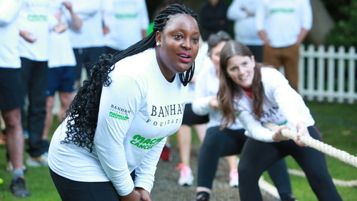A woman with long braids is leaning over and looking at something. She is wearing a long sleeved white t-shirt and pink lipstick. Behind her are a group of people pulling at a rope. 