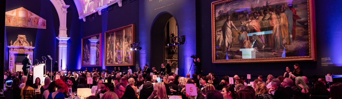 A view of an art gallery where an event is being hosted. Across the hall are tables with people sitting. They are dress in formal attire.