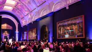 A view of an art gallery where an event is being hosted. Across the hall are tables with people sitting. They are dress in formal attire.