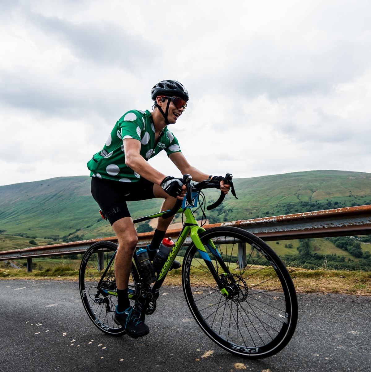 A man in a green Macmillan top is road biking up a hill.