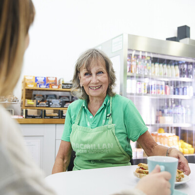 Jenny is sat at a table in the Macmillan Horizon Centre's café. She is wearing a green polo shirt and a green apron with the Macmillan logo on it. She appears to be talking to someone.